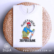 Футболка Найкращий зварювальник, білого кольору, зображена на столі з верхнього ракурсу, фон світло-сірий, принт з малюнком зварювальника у синій куртці та червоній касці, що працює з електродом, текст на футболці чорний, футболка лежить на плетеній основі.