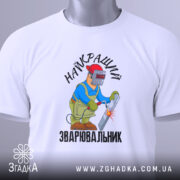Футболка Найкращий зварювальник, білого кольору, зображена на столі з верхнього ракурсу, фон світло-сірий, принт з малюнком зварювальника у синій куртці та червоній касці, що працює з електродом, текст на футболці чорний, на футболці є етикетка.