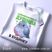Футболка найприскіпливіша душніла білого кольору, складена на поверхні, фон світло-сірий, принт з малюнком пташки та текстом зеленого кольору, на футболці є етикетка з зеленим шнурком.