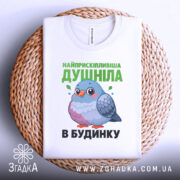 Футболка найприскіпливіша душніла білого кольору, складена на плетеному кошику, фон світлий, принт з малюнком пташки та текстом зеленого кольору.