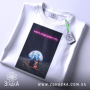 Футболка нам є куди дивитись. Біла футболка, складена на поверхні, з принтом, на якому видно двох людей, що сидять на лавці під землею, оточеними зоряним небом. Текст на принті яскравий, неоновий.