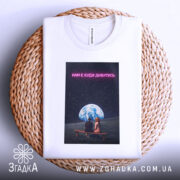 Футболка нам є куди дивитись. Біла футболка, складена на плетеній основі, з принтом, де видно двох людей, що сидять під землею на фоні зоряного неба. Текст на принті виконаний у яскравому неоновому стилі.
