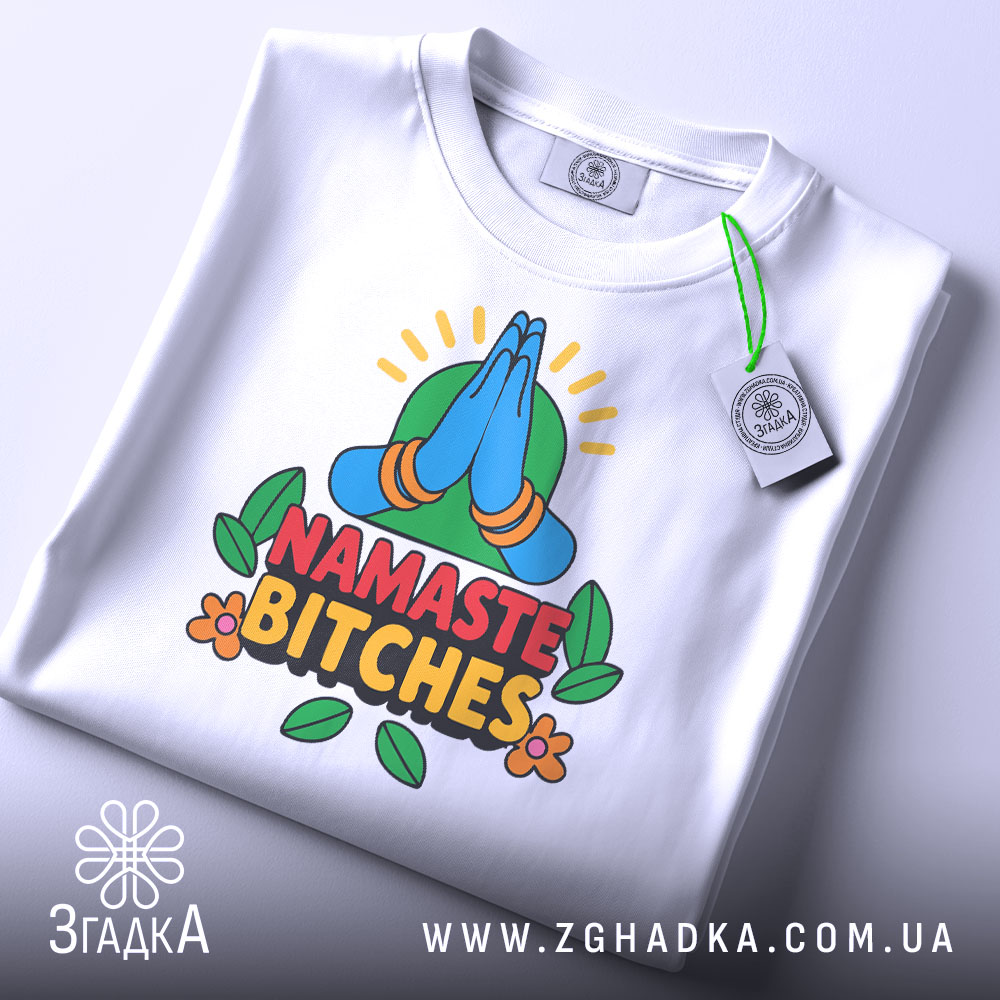 Футболка намасте бічес, біла, зображена зверху, складена на поверхні, фон світло-сірий, принт з зображенням складених рук у позі намасте, кольорові деталі з зеленим фоном та написом 'NAMASTE BITCHES' у червоному та чорному кольорах, на футболці є етикетка.