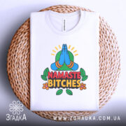 Футболка намасте бічес, біла, зображена зверху, складена на плетеній кошику, фон світло-сірий, принт з зображенням складених рук у позі намасте, кольорові деталі з зеленим фоном та написом 'NAMASTE BITCHES' у червоному та чорному кольорах.