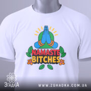 Футболка намасте бічес, біла, зображена зверху, складена на поверхні, фон світло-сірий, принт з зображенням складених рук у позі намасте, кольорові деталі з зеленим фоном та написом 'NAMASTE BITCHES' у червоному та чорному кольорах, на футболці є етикетка.