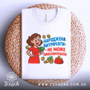 Футболка народжена витрачати, біла, зображена зверху на плетеному кошику, фон білий, принт з жінкою в червоній сукні, яка тримає гроші, текст яскравий, синій та червоний, футболка акуратно складена.