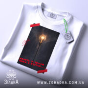 Футболка Навіть у темні часи є світло, біла футболка складена на поверхні, фон білий, принт з лампою на фоні темного міста, червона стрічка на принті, етикетка з зеленим шнурком.