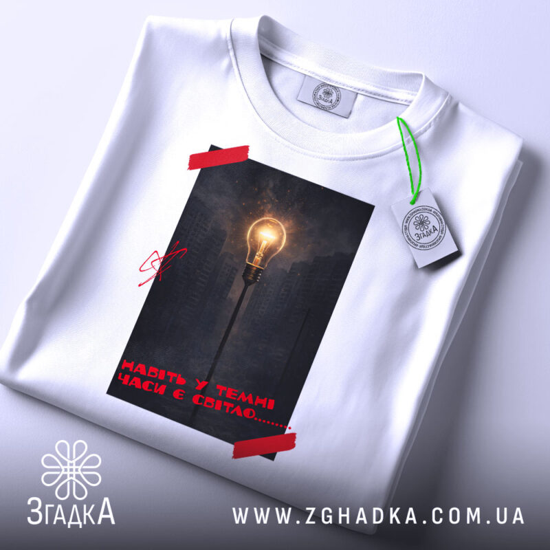 Футболка Навіть у темні часи є світло, біла футболка складена на поверхні, фон білий, принт з лампою на фоні темного міста, червона стрічка на принті, етикетка з зеленим шнурком.