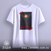 Футболка Навіть у темні часи є світло, біла футболка на вішалці, фон світло-сірий, принт з лампою на фоні темного міста, червона стрічка на принті.