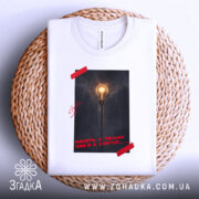 Футболка Навіть у темні часи є світло, біла футболка складена на кошику, фон білий, принт з лампою на фоні темного міста, червона стрічка на принті.