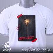 Футболка Навіть у темні часи є світло, біла футболка складена на поверхні, фон білий, принт з лампою на фоні темного міста, червона стрічка на принті, етикетка з зеленим шнурком.