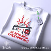 Футболка не бути електриком. Біла футболка, що лежить на столі, з принтом червоного кольору, зображення електричного шнура та написом "НЕ БУТИ МЕНІ ЕЛЕКТРИКОМ!". На футболці є етикетка з зеленим шнурком. Фон світло-сірий.