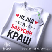 Футболка не дід а бабусин краш. Біла футболка, складена на плоскій поверхні, з видимим етикеткою. Принт на футболці чорного та червоного кольорів з текстом 'НЕ ДІД А БАБУСИН КРАШ'. Фон світло-сірий, що створює контраст з футболкою.