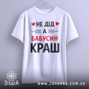 Футболка не дід а бабусин краш. Біла футболка з коротким рукавом, підвішена на вішалці, зображена з фронтального ракурсу. Принт чорного та червоного кольорів з текстом 'НЕ ДІД А БАБУСИН КРАШ'. Фон світло-сірий, що підкреслює чистоту дизайну.