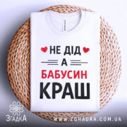 Футболка не дід а бабусин краш. Біла футболка, складена на круглій плетеній підставці. Принт чорного та червоного кольорів з текстом 'НЕ ДІД А БАБУСИН КРАШ'. Фон світло-сірий, що надає елегантності загальному вигляду.