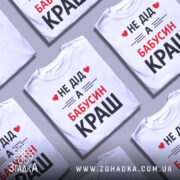 Футболка не дід а бабусин краш. Біла футболка з коротким рукавом, зображена з фронтального ракурсу на манекені. На футболці великий принт чорного та червоного кольорів з текстом 'НЕ ДІД А БАБУСИН КРАШ'. Фон світло-сірий, що підкреслює яскравість принта.