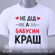 Футболка не дід а бабусин краш. Біла футболка, складена на плоскій поверхні, з видимим етикеткою. Принт на футболці чорного та червоного кольорів з текстом 'НЕ ДІД А БАБУСИН КРАШ'. Фон світло-сірий, що створює контраст з футболкою.