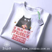 Футболка не люблю працювати, білого кольору, зображена зверху на столі, фон світло-сірий, принт з котом, що тримає шматок їжі, з текстом червоного кольору, на комірці є етикетка.