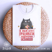 Футболка не люблю працювати, білого кольору, зображена зверху на плетеному кошику, фон світло-сірий, принт з котом, що тримає шматок їжі, з текстом червоного кольору.