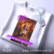 Футболка не на то нарвались. Біла футболка, складена на поверхні, з принтом дівчини в коричневій сукні, фон з вогнем. На футболці є етикетка з зеленим шнурком.