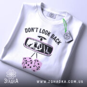 Футболка не озирайся назад. Біла футболка, складена на поверхні, з видимим принтом. На футболці напис 'DON'T LOOK BACK' та малюнок дзеркала заднього виду з черепом і кістками. Фон світло-сірий, а на футболці видно етикетку з зеленим шнурком.