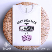 Футболка не озирайся назад. Біла футболка, складена на плетеній основі, з видимим принтом. На футболці напис 'DON'T LOOK BACK' та малюнок дзеркала заднього виду з черепом і кістками. Фон білий, а плетена основа додає текстури до зображення.