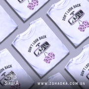 Футболка не озирайся назад. Біла футболка з коротким рукавом, зображена з фронтального ракурсу на манекені. На футболці принт з написом 'DON'T LOOK BACK' та малюнком дзеркала заднього виду, з якого видно череп і дві кістки. Фон світло-сірий, що підкреслює яскравість принта.