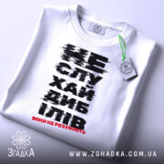 Футболка не слухай дебілів, біла, зображена зверху на плоскій поверхні, фон світлий, принт чорного кольору з червоним акцентом, на футболці є етикетка з зеленим шнурком.