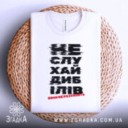 Футболка не слухай дебілів, біла, зображена зверху на плоскій поверхні з плетеного матеріалу, фон світлий, принт чорного кольору з червоним акцентом, текст розмитий.