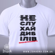 Футболка не слухай дебілів, біла, зображена зверху на плоскій поверхні, фон світлий, принт чорного кольору з червоним акцентом, на футболці є етикетка з зеленим шнурком.