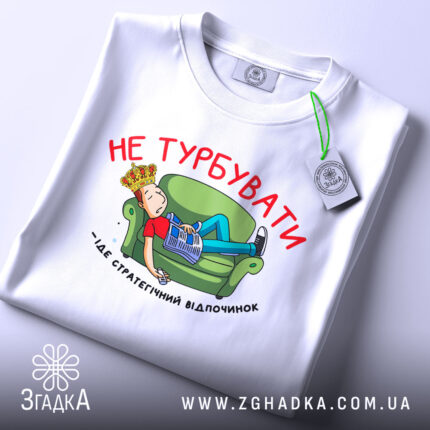 Футболка не турбувати, біла, з принтом на грудях, зображення хлопця на зеленому дивані, газета в руках, текст червоним кольором, футболка складена на поверхні.