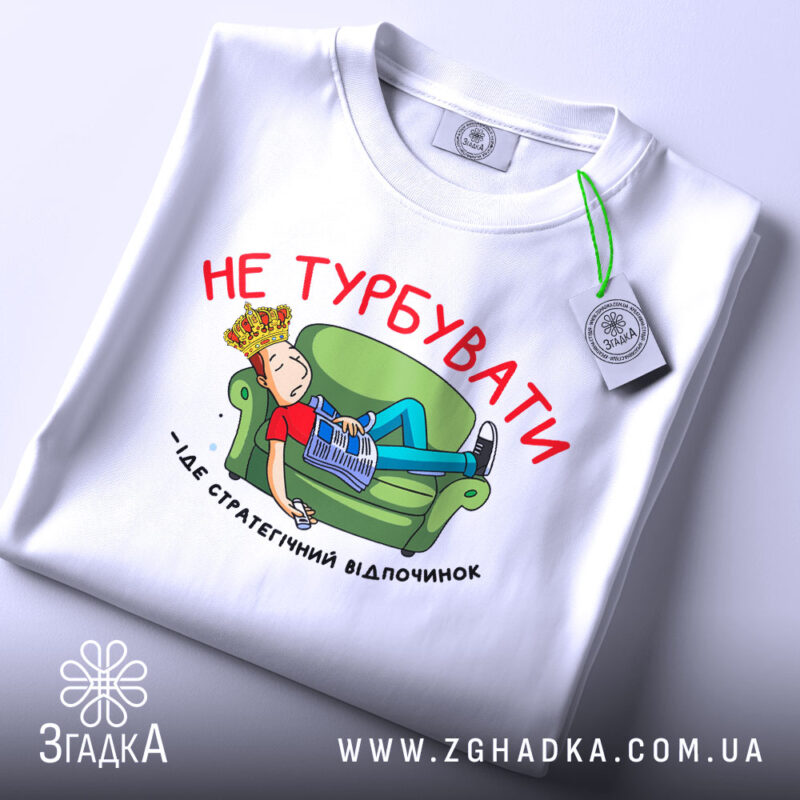 Футболка не турбувати, біла, з принтом на грудях, зображення хлопця на зеленому дивані, газета в руках, текст червоним кольором, футболка складена на поверхні.