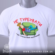 Футболка не турбувати, біла, з принтом на грудях, зображення хлопця на зеленому дивані, газета в руках, текст червоним кольором, футболка складена на поверхні.