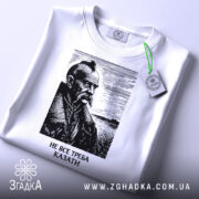 Футболка не все треба казати, біла, складена на поверхні, фон світлий, принт з портретом чоловіка з вусами та бородою, напис під портретом: 'НЕ ВСЕ ТРЕБА КАЗАТИ', на етикетці зелена стрічка.