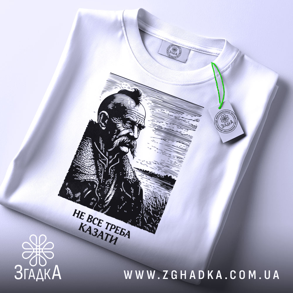 Футболка не все треба казати, біла, складена на поверхні, фон світлий, принт з портретом чоловіка з вусами та бородою, напис під портретом: 'НЕ ВСЕ ТРЕБА КАЗАТИ', на етикетці зелена стрічка.