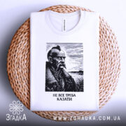 Футболка не все треба казати, біла, складена на плетеній основі, фон світлий, принт з портретом чоловіка з вусами та бородою, напис під портретом: 'НЕ ВСЕ ТРЕБА КАЗАТИ'.