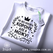 Футболка не всі носять корону. Біла футболка, складена на поверхні, з чорним принтом, що містить текст 'НЕ ВСІ НОСЯТЬ КОРОНУ, АЛЕ Я МОЖУ!'. На футболці є етикетка з зеленим шнурком. Фон світло-сірий.