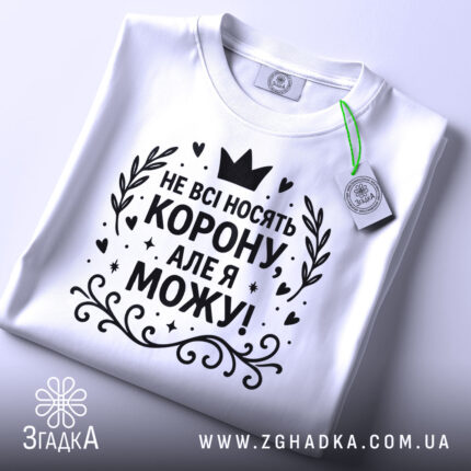 Футболка не всі носять корону. Біла футболка, складена на поверхні, з чорним принтом, що містить текст 'НЕ ВСІ НОСЯТЬ КОРОНУ, АЛЕ Я МОЖУ!'. На футболці є етикетка з зеленим шнурком. Фон світло-сірий.