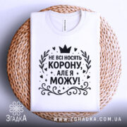 Футболка не всі носять корону. Біла футболка, розкладена на плетеній кошику, з чорним принтом, що містить текст 'НЕ ВСІ НОСЯТЬ КОРОНУ, АЛЕ Я МОЖУ!'. Фон світло-сірий.