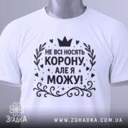 Футболка не всі носять корону. Біла футболка, складена на поверхні, з чорним принтом, що містить текст 'НЕ ВСІ НОСЯТЬ КОРОНУ, АЛЕ Я МОЖУ!'. На футболці є етикетка з зеленим шнурком. Фон світло-сірий.