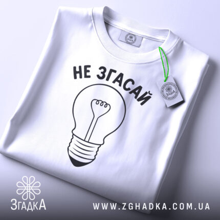 Футболка не згасай, біла, складена на поверхні, фон світло-сірий, принт у вигляді лампочки з написом 'НЕ ЗГАСАЙ' чорного кольору, з етикеткою зеленого кольору.