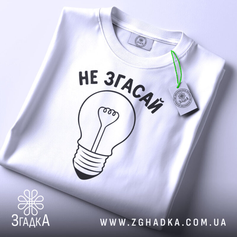 Футболка не згасай, біла, складена на поверхні, фон світло-сірий, принт у вигляді лампочки з написом 'НЕ ЗГАСАЙ' чорного кольору, з етикеткою зеленого кольору.