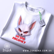 Футболка не зли лисичку. Біла футболка, складена на поверхні, з принтом лисички, що має червоні очі. На футболці є етикетка з зеленим шнурком, фон світлий, що підкреслює деталі принта.