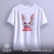 Футболка не зли лисичку. Біла футболка, підвішена на вішалці, з принтом лисички, що має червоні очі. Фон світлий, з легким градієнтом, що створює акцент на футболці.