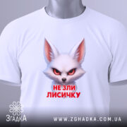 Футболка не зли лисичку. Біла футболка, складена на поверхні, з принтом лисички, що має червоні очі. На футболці є етикетка з зеленим шнурком, фон світлий, що підкреслює деталі принта.