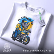 Футболка не зупиняйся. Біла футболка, складена на поверхні, з принтом носорога в жовтій куртці на мотоциклі, фон світло-сірий, на футболці є етикетка з зеленим шнурком.