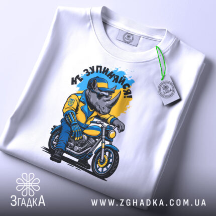 Футболка не зупиняйся. Біла футболка, складена на поверхні, з принтом носорога в жовтій куртці на мотоциклі, фон світло-сірий, на футболці є етикетка з зеленим шнурком.
