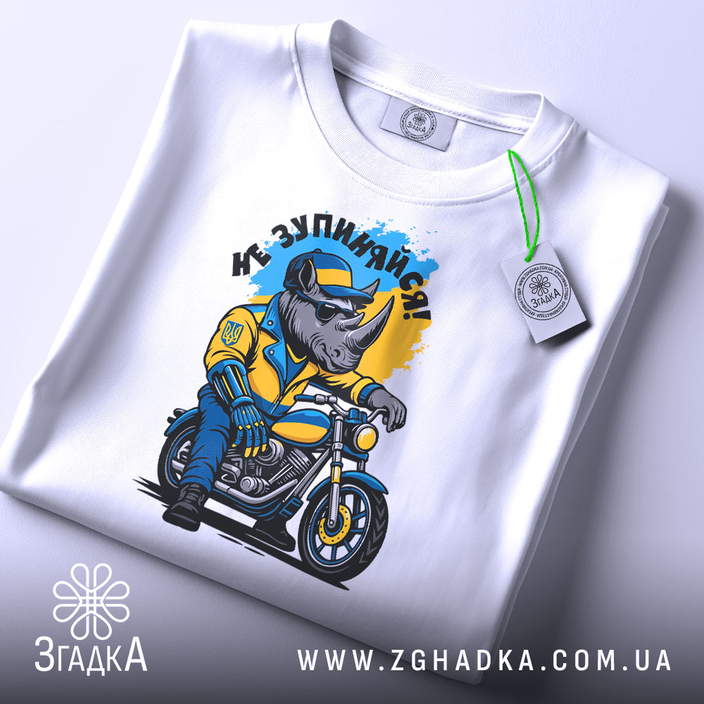 Футболка не зупиняйся. Біла футболка, складена на поверхні, з принтом носорога в жовтій куртці на мотоциклі, фон світло-сірий, на футболці є етикетка з зеленим шнурком.