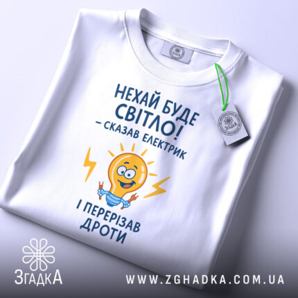 Футболка нехай буде світло. Біла футболка, складена на поверхні, ракурс зверху, фон світлий, принт з жовтою лампочкою та текстом у синьому кольорі, з зеленим ярликом.