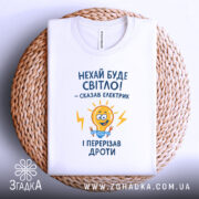 Футболка нехай буде світло. Біла футболка, складена на плетеній основі, ракурс зверху, фон натуральний, принт з жовтою лампочкою та текстом у синьому кольорі.